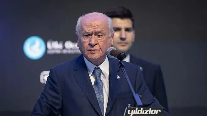 Bahçeli kürsüde lacıvert takım elbisesiyle ayakta konuşma yapıyor. Beyaz seyrek saçları ile sert bir duruşu var. Arkada bir erkek daha duruyor.