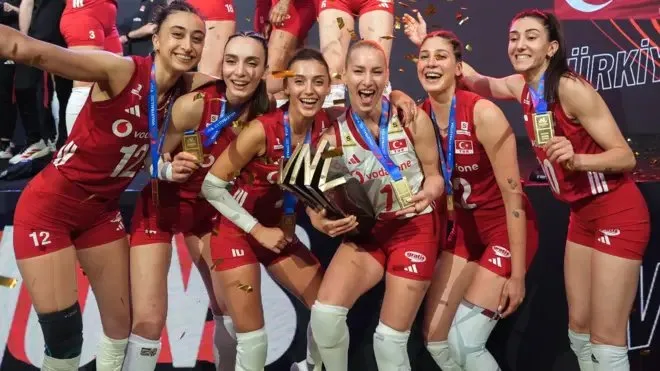 Voleybol milli takımı oyuncuları podyumda kupayı kaldırırken gülümsüyor, havada konfetiler uçuşuyor