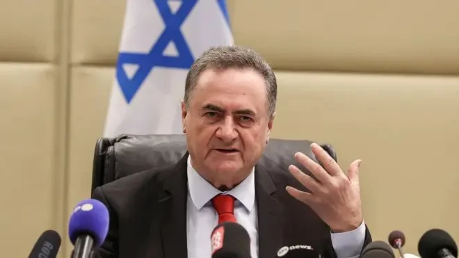 Israel Katz üzerinde bir takım elbiseyle bir masada oturmuş, yüzünde gergin bir ifadeyle konuşurken elini kaldırıyor. Önünde mikrofonlar, arkasında ise bir İsrail bayrağı var