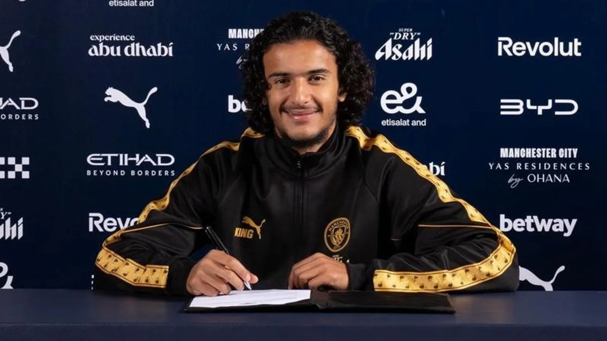 Ayyoub Bouaddi in a Manchester City tracksuit top holding a pen with a contract on the desk in front of him