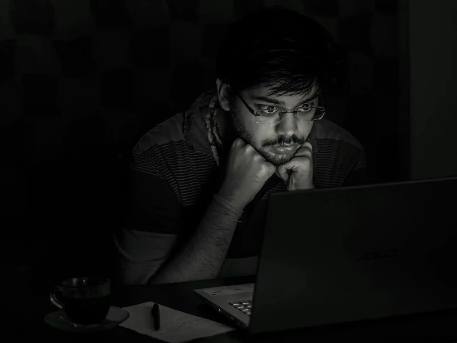 Black and white photo of a man in dark room, his face illuminated by the glow of a computer screen