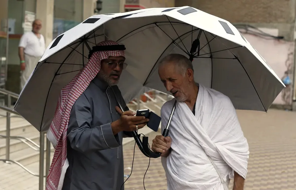 Two men stand under an umbrella as one shows the other something on a smartphone.