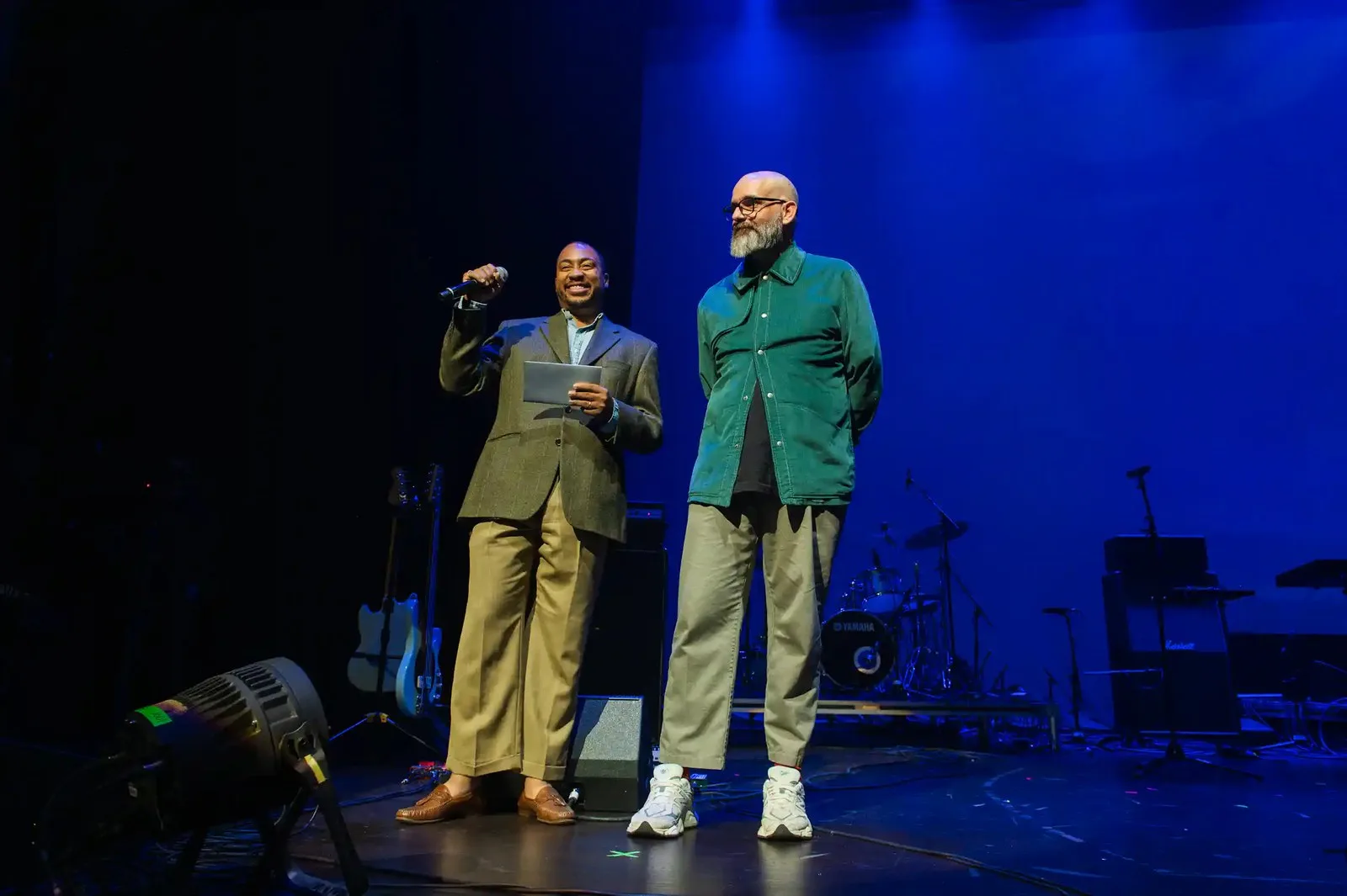 Two men stand on a stage and one holds a microphone.